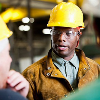 Three men wearing yellow hard hats talking to one another.  The backs of two of the men are turned to the camera and the focus is on the man facing the camera.  The man facing forward is black, in his twenties and wearing protective eyeglasses and a brown coat.